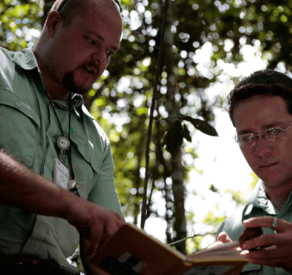 Two Vale employees are out in the open, with trees in the background. One of them is holding a notebook and both are looking toward the object.