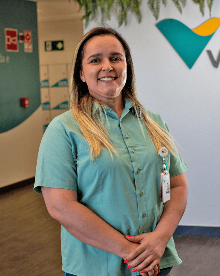 Mulher parada em um espaço de escritório, usando camisa verde clara. Ela está sorrindo e com as mãos cruzadas a frente do corpo.