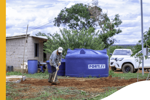 In a dirt area, a man weeds the soil with an ax. In the background, it is possible to see two cars, a water tank and a small house, in addition to trees.