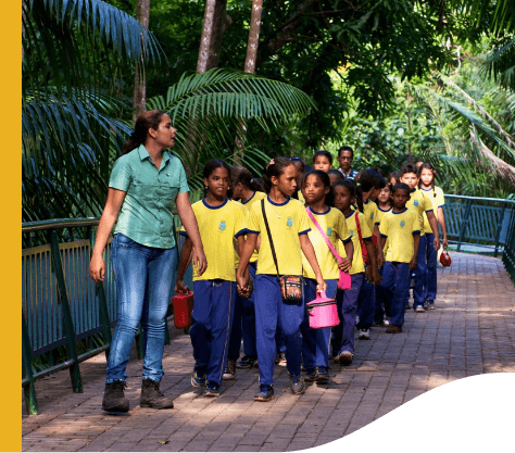 Uma empregada Vale acompanha um grande grupo de crianças, todas vestidas com roupas escolares, por um caminho arborizado.