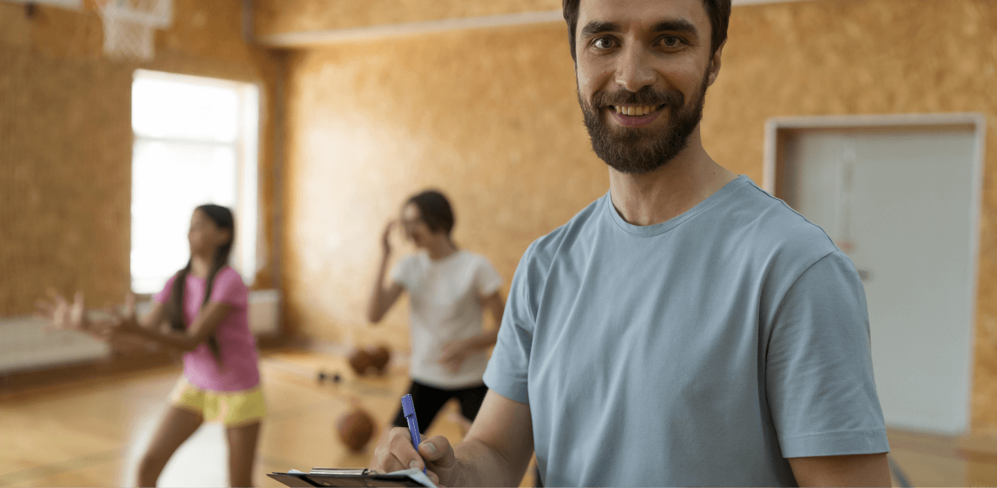 Man smiling at camera, holding a clipboard and a pen. He has a beard and short, straight, brown hair. Behind him, there are two people doing physical exercises.