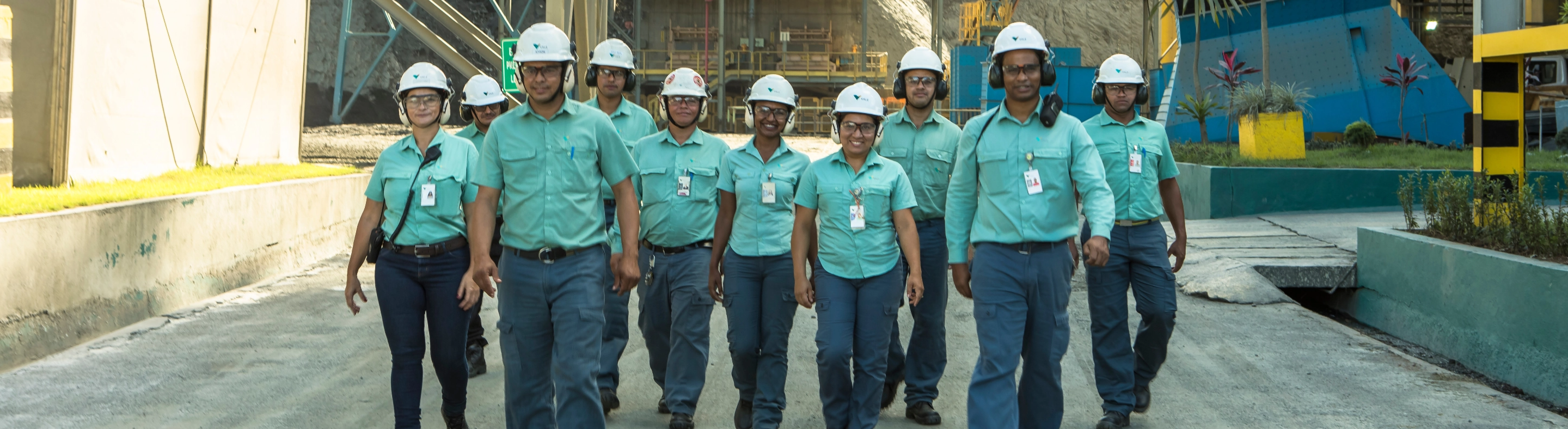 Photo of three men and a woman talking in an operation area with mountains of dirt and a machine in the background. The four are wearing uniforms, green button-down shirts, white helmets with Vale logo, goggles and ear protection.