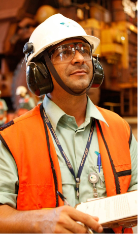 Belly to head shot of a white man with an operation in the background, wearing a Vale uniform: green button-up shirt, orange vest, goggles, helmet, ear protection and badge, holding a pen and clipboard