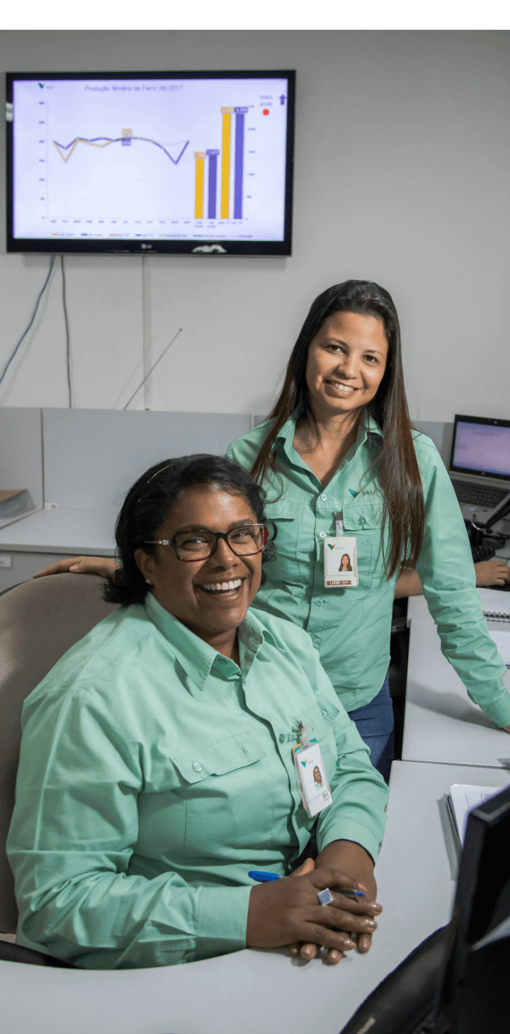 Foto de duas mulheres, uma delas está sentada usando óculos e tem cabelo curto, a outra está em pé ao lado, sorrindo e possui cabelo longo. As duas estão usando o uniforme verde da Vale.