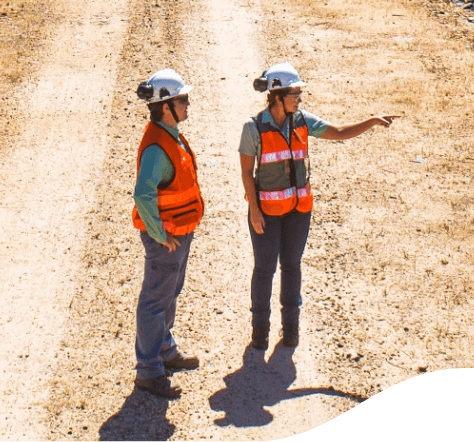 Foto vista de cima, do corpo odo de dois empregados da Vale, conversando em um local de terra e areia, utilizando uniforme, calça cinza, camisa Verde, colete laranja, capacete e óculos.