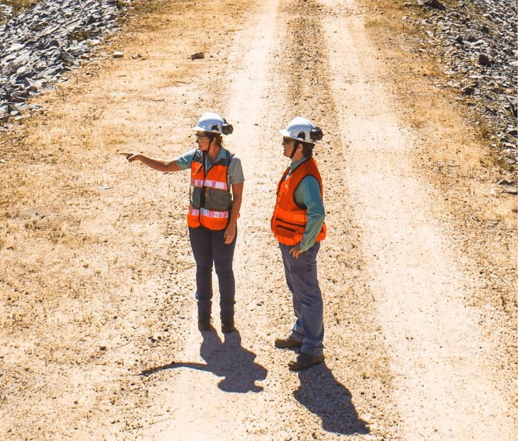 Full body photo of 10 Vale employees with an operation in the background, wearing uniforms: pants, green button-up shirt, helmet, goggles, ear protection and badge