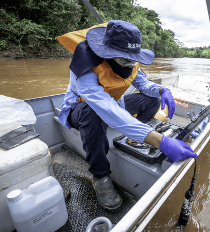 Um homem, de chapéu, máscara, óculos protetor, luvas e colete, está dentro de um barco com diversos galões e uma maleta com equipamentos.