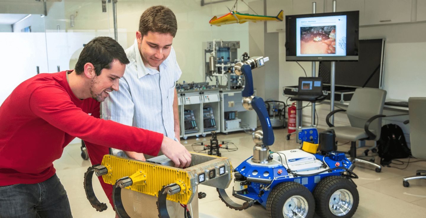 Two young men are working on iron equipment. Both are standing and smiling.