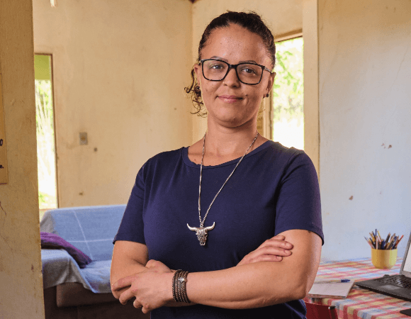 Retrato de Roziane Duarte sorrindo para a câmera. Ela está com braços cruzados, usa óculos, um colar grande e está com o cabelo amarrado.