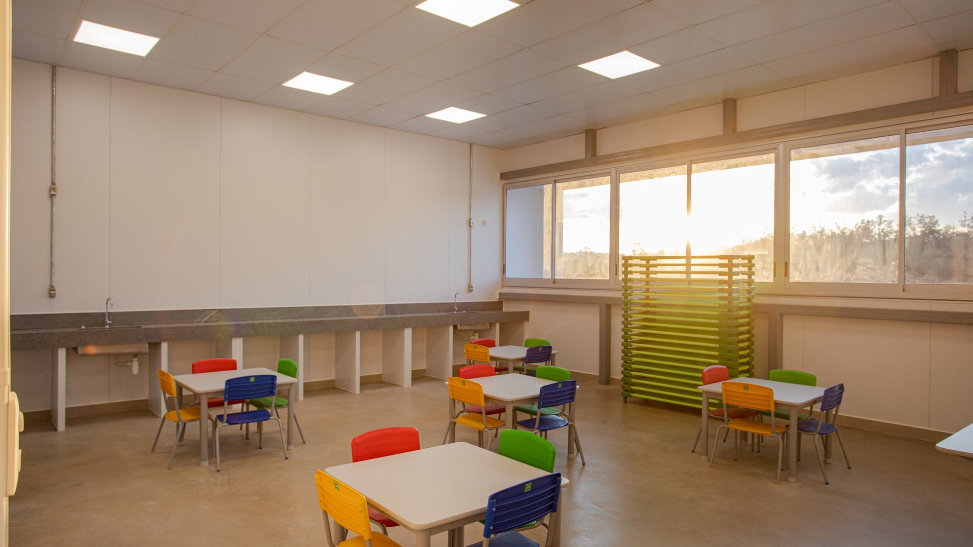 Uma sala com janelas amplas e diversas mesas com cadeiras coloridas ao redor. Ao fundo há uma grande bancada com duas pias.