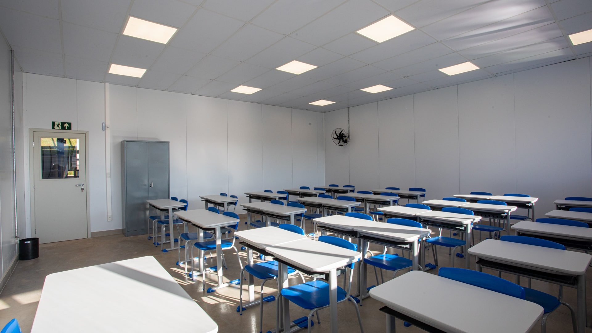 Diversas mesas e cadeiras dispostas lado a lado em uma sala de aula. Há um pequeno armário e um ventilador na parede.