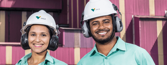 Imagem de dois empregados Vale, uma mulher e um homem. Os dois estão sorrindo para foto e usam capacete de proteção, além do uniforme verde da empresa. Ao fundo há containers roxos.