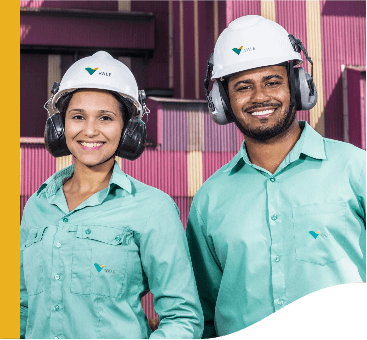 Imagem de dois empregados Vale, uma mulher e um homem. Os dois estão sorrindo para foto e usam capacete de proteção, além do uniforme verde da empresa. Ao fundo há containers roxos.