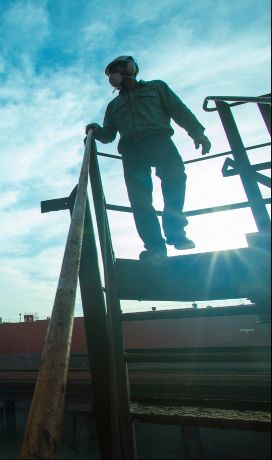 Homem descendo uma escada em um espaço de operações. Ele está contra o sol e a foto está sombreada. Ao fundo há um céu azul.