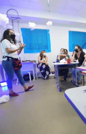 A woman is standing in a classroom while other women are sitting looking at her. All are wearing masks.