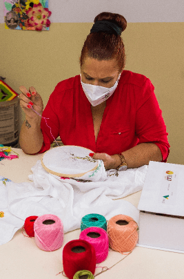 A woman is seated, embroidering. There are several colorful threads in front of her.