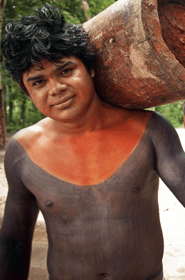 An indigenous person with his body painted black and red. He holds a piece of a tree trunk on his shoulders.