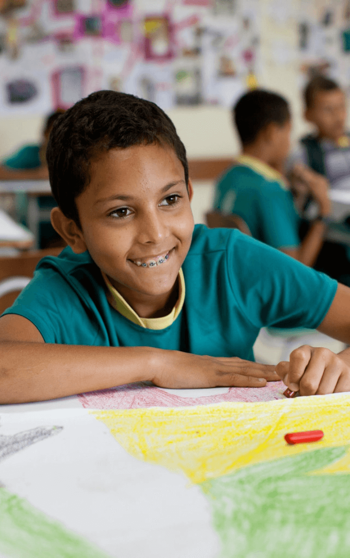 Um menino, dentro de uma sala de aula, pinta uma folha com giz de cera. Ele usa aparelho nos dentes e veste um uniforme verde e amarelo.