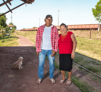 Imagem de um senhor e uma senhora. Os dois estão de pé, sorrindo para foto. Ao fundo é possível ver uma área gramada e a um trem.