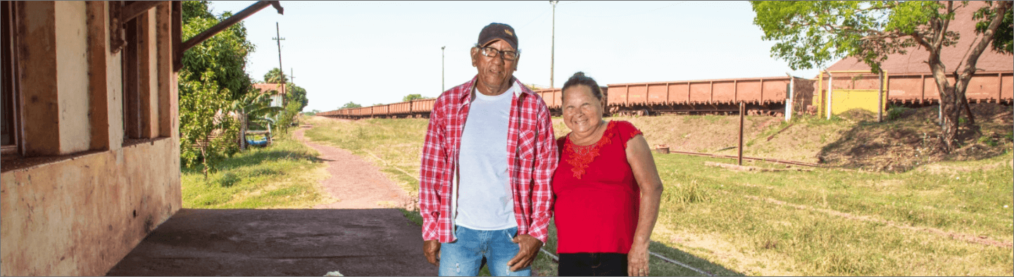 Imagem de um senhor e uma senhora. Os dois estão de pé, sorrindo para foto. Ao fundo é possível ver uma área gramada e a um trem.