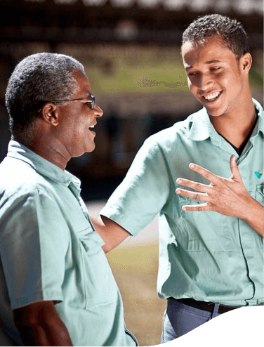 Dois empregados Vale, um senhor e um jovem, se olham e sorriem. O jovem está com uma mão no ombro do senhor e gesticula com a outra. Ambos estão com uma camisa verde de uniforme.