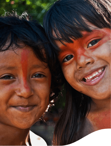 Duas crianças indígenas sorrirem lado a lado para foto. Ambas estão com pinturas vermelhas no rosto.