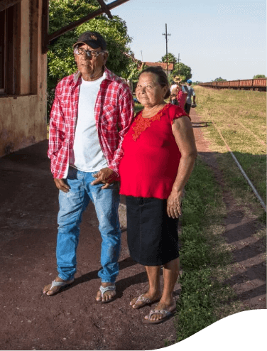 Um senhor e uma senhora estão de pé em um local gramado, em que é possível ver um trem ao fundo. Ele usa calça jeans, camiseta branca e camisa xadrez vermelha, além de óculos e boné. Ela usa uma saia preta e uma blusa vermelha. Ambos estão de chinelo.