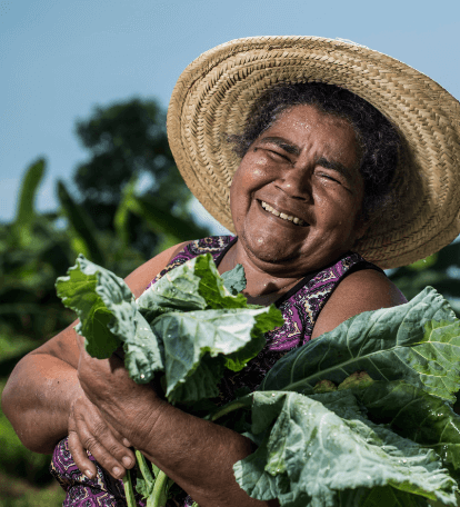 Uma senhora segura verduras nos braços. Ela está sorrindo e usa um chapéu de palha.