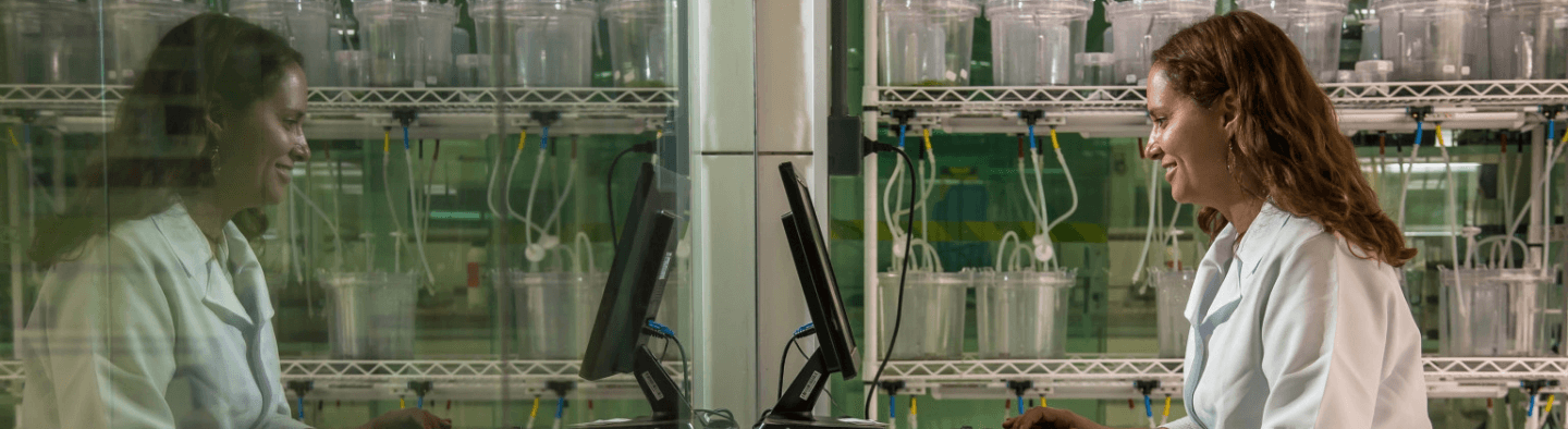 Uma cientista, de jaleco branco, olha para tela de um computador e sorri. Ao fundo é possível ver alguns recipientes transparentes com fios conectados a eles.