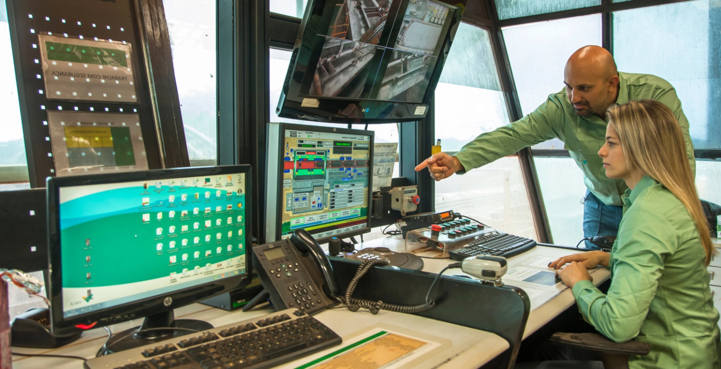 Two Vale employees, a bald man with a beard and a blond woman with straight hair, are in a control room, full of monitors. The man is standing, pointing at a screen and the woman is sitting, paying attention.