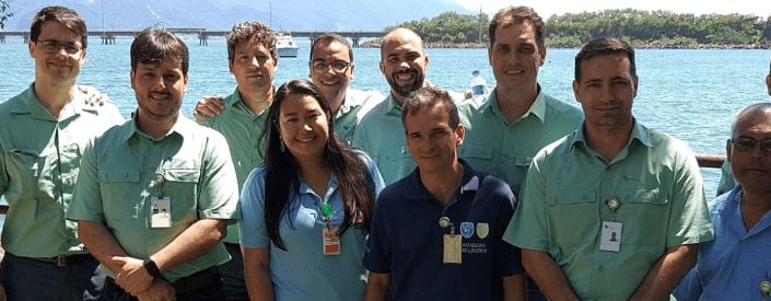 Photo of eight men and one woman with sea and a bridge in the background on a sunny day. Seven men are wearing Vale uniforms, short-sleeved green button-down shirts, jeans and badges. One of the men is wearing a navy blue polo shirt, jeans and a badge. The woman is wearing a light blue polo shirt, jeans and a badge.