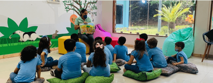 Enquanto uma empregada Vale mostra um livro, algumas crianças olham em direção a ela. As crianças estão sentadas em almofadas e usam uniformes azuis.