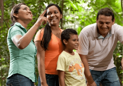 Em um ambiente ao ar livre, uma empregada Vale aponta para cima. Ao lado dela, há uma mulher, um menino e um homem, todos sorriem.