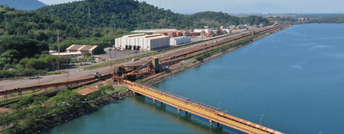 Imagem aérea do porto de Itaguaí. É possível ver uma ponte sob a água e, ao fundo, alguns galpões e uma área montanhosa.