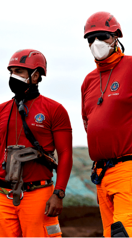 Dois bombeiros de pé, ambos de capacete e máscara de proteção.