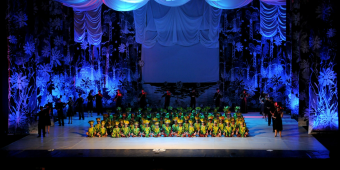 Diversas crianças sentadas no palco de teatro com fantasias iguais e ao redor alguns músicos clássicos 
