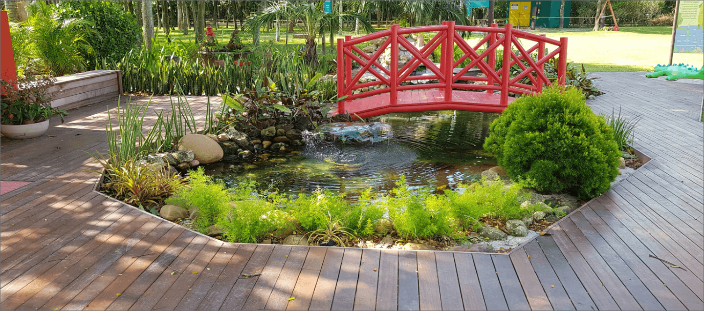 Imagem de um jardim com uma ponte de madeira vermelha em cima de um pequeno lago