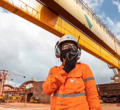 Photo of a woman in an operation area talking on a radio. She is wearing a Vale uniform, orange shirt, black glove, face mask, goggles, white helmet with Vale logo, and ear protection.