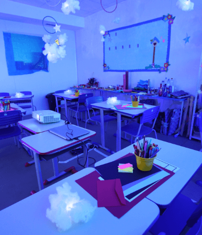 Sala de aula com a luz apagada e alguns pontos de luz espalhados pelas mesas, onde também há papeis e lápis coloridos.
