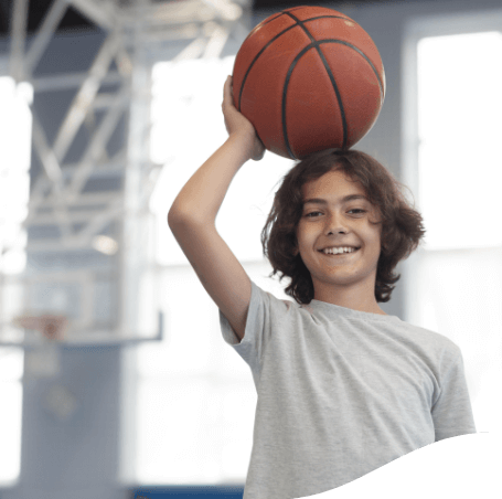 Photo of a boy smiling at camera while holding a basketball ball over his head. He has medium-long, straight, brown hair and is on a sports court.