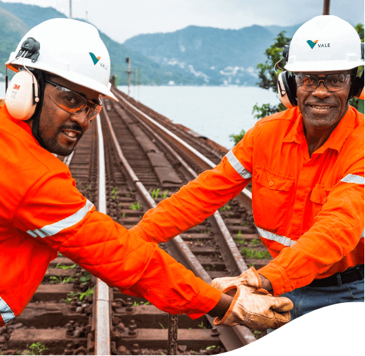 Dois homens negros com uniformes laranjas, capacetes e óculos de proteção, trabalhando em um trilho de ferrovia.