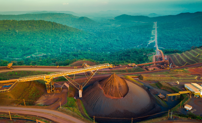 Imagem aérea de uma mina a céu aberto, mostrando uma grande pilha de minério cercada por maquinário industrial e estradas. Ao fundo, há uma vasta área de floresta verdejante e montanhas ao longe. 