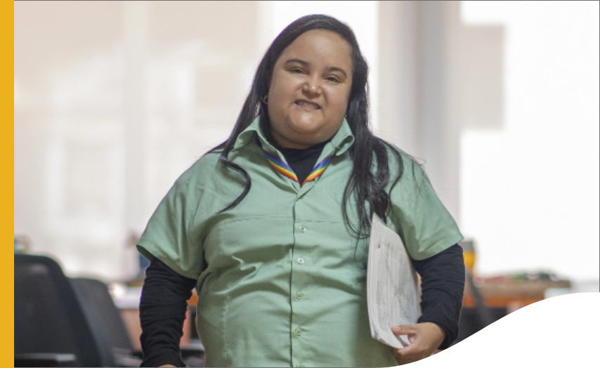 Uma mulher está segurando uma pasta embaixo do braço e sorrindo para foto. Ela está com uniforme Vale.