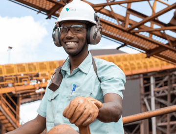 In an outdoor operation area, a Vale employee is smiling for the photo. He is wearing goggles, a helmet and gloves. On his shoulder, there is a radio transmitter fixed.