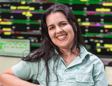 Foto de uma mulher em um escritório e vários monitores ao fundo. Ela está apoiada em um balcão e está usando camisa verde de botões com logo da Vale, cabelo comprido solto liso e está sorrindo.