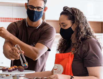 Um homem e uma mulher, ambos de camiseta marrom e máscara de proteção, estão em uma cozinha manuseando brigadeiros.
