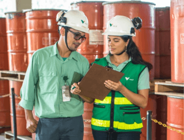 Um homem e uma mulher parados lado a lado olhando uma prancheta. Eles usam calças jeans, blusas verdes de uniforme e capacetes de proteção com o logotipo da Vale. A mulher usa um colete verde escuro, com detalhes em verde limão. Ao fundo estão vários contêineres empilhados.