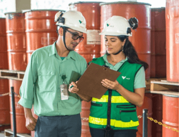 Um homem e uma mulher parados lado a lado olhando uma prancheta. Eles usam calças jeans, blusas verdes de uniforme e capacetes de proteção com o logotipo da Vale. A mulher usa um colete verde escuro, com detalhes em verde limão. Ao fundo estão vários contêineres empilhados.