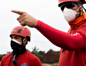 Dois bombeiros estão olhando para o horizonte e, um deles, aponta para frente. Eles estão usando máscara de proteção e capacete.