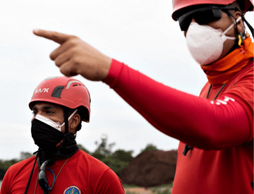 Dois bombeiros estão olhando para o horizonte e, um deles, aponta para frente. Eles estão usando máscara de proteção e capacete.